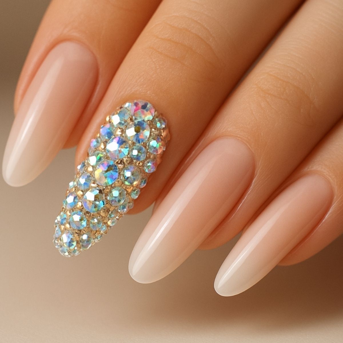 A hand with long, almond-shaped nails, four painted in a nude shade and one accent nail featuring shimmering Crystal Nail Art with shiny, multi-colored rhinestones.