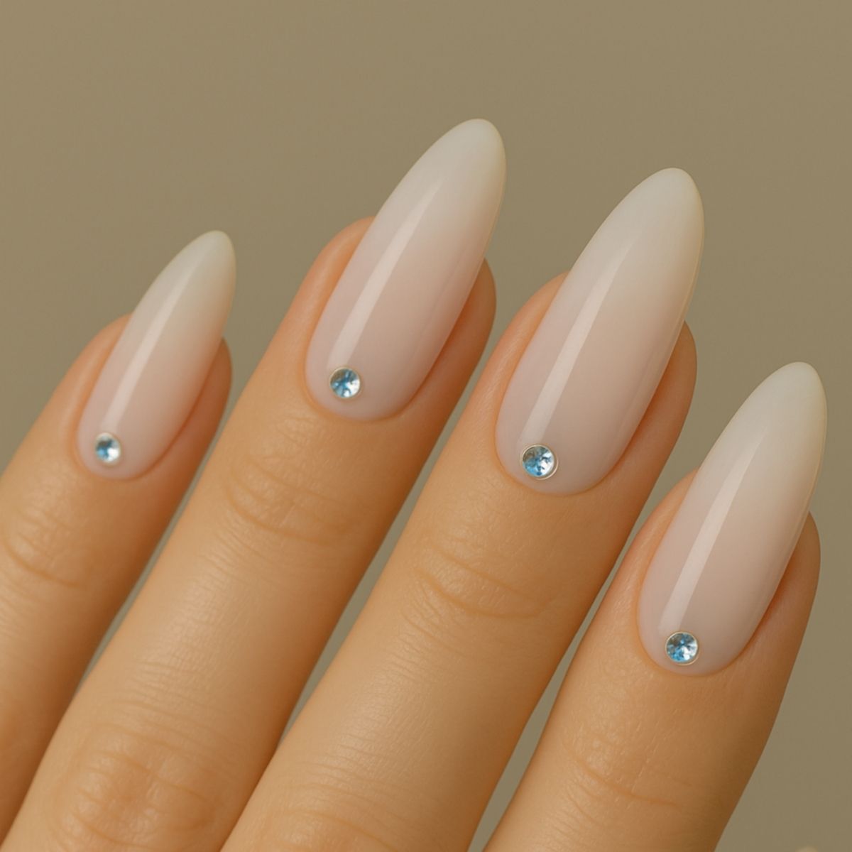 A hand with almond-shaped nude nails, each adorned with a single small blue Swarovski crystal near the cuticle, showcases elegant Crystal Nail Art against a neutral background.