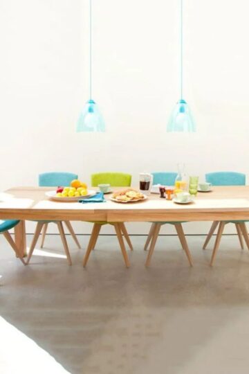 A wooden extendable dining table with six colorful chairs, breakfast foods, drinks, and fruit on top sits under two blue pendant lights in a bright room—perfect for modern interior design with impact.