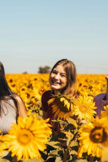 Three women standing and smiling among blooming sunflowers in a field on a sunny day, radiating joy and happiness.