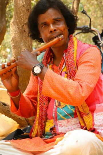 A man in colorful traditional attire plays Indian Classical Flute Music outdoors, seated on the ground near trees and a parked motorcycle, filling the air with serene stillness.