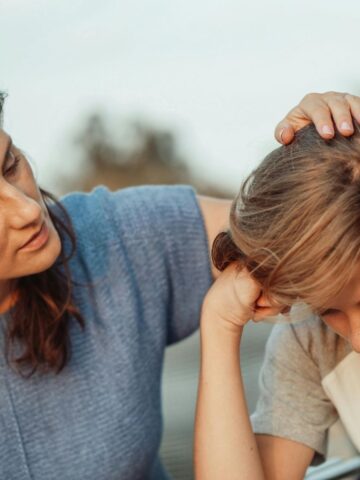 A woman comforts a young person who appears upset, gently placing her hand on their head while talking to them outdoors, soothing child worries with compassion and care.