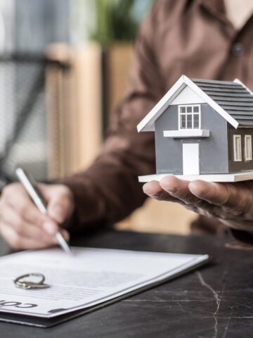 A person holds a small model house in one hand while signing a document, perhaps weighing the difference between selling for cash or listing with an agent on a clipboard.