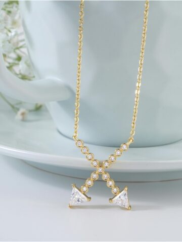 A gold necklace with an X-shaped pendant adorned with round and triangular clear gemstones, elegantly displayed in front of a white cup and saucer-a classic gift, perfect for lovers of gold diamond necklaces.
