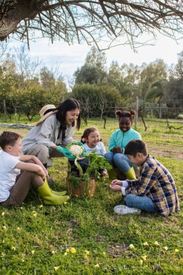 An adult and four children sit under a tree in a grassy area, gathered around potted plants, enjoying eco-friendly activities as they garden and observe nature together.