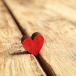 A small red wooden heart stands upright on a light-colored wooden surface with visible grain and cracks, symbolizing manifesting love and setting intentions for meaningful connections.