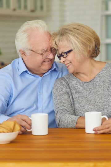 An elderly couple sits at a wooden table in a cozy home environment, smiling and leaning towards each other, each holding a white mug, with a plate of cookies on the table.