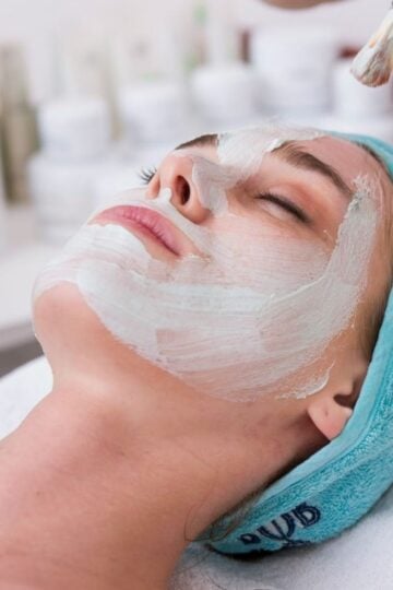 A person lies down with eyes closed, enjoying a cosmetic treatment as a brush applies a white mask to their face; they wear a blue headband—one of many relaxing cosmetic procedures.