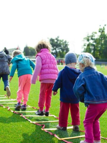 Several young children walk in a line on a sports field, stepping through a ladder laid flat on the grass during an outdoor activity that encourages child development.
