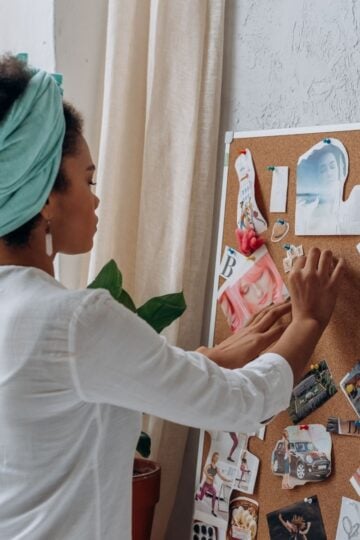 A woman stands by a corkboard, arranging various photos, magazine cutouts, and notes pinned on her Recovery Wall in a well-lit room.