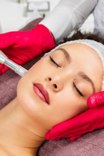 A person lies with eyes closed as a professional wearing red gloves performs a facial treatment using a handheld device.