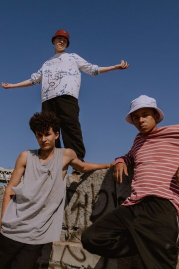 Three young men pose on a graffiti-covered concrete structure, wearing casual clothes and bucket hats, with a vape in hand and pills scattered nearby, against a clear blue sky.