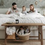 A family of three sits on a bed in a cozy bedroom, creating the perfect sleep environment. In the foreground, a wooden bench holds a basket with teddy bears and a wicker basket with a white blanket.