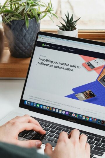 A person types on a laptop displaying the Shopify website, which shows a banner about starting an online store; potted plants are in the background.