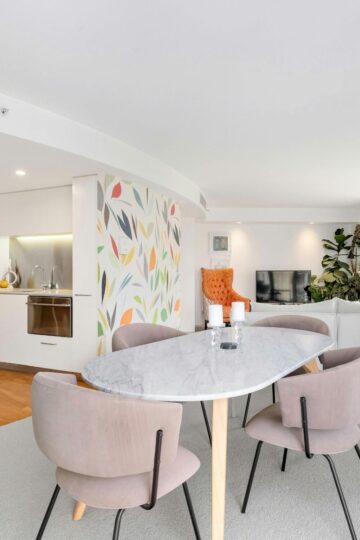 Modern open-plan living area featuring thoughtful interior design with a marble dining table, four upholstered chairs, small kitchen, abstract wall art, and bright natural light streaming through large windows.