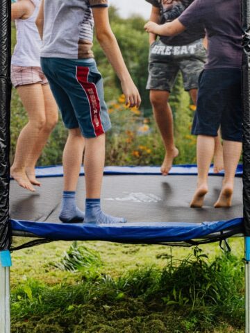 Four children are jumping together on an outdoor trampoline in a grassy yard, making it one of the best kids activities and active day out ideas that's fun for parents too.