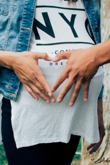 Two people form a heart shape with their hands over the belly of a pregnant person—wearing a "NYC" t-shirt and denim jacket—who is standing outdoors, joyfully embracing parenthood on their own timeline.
