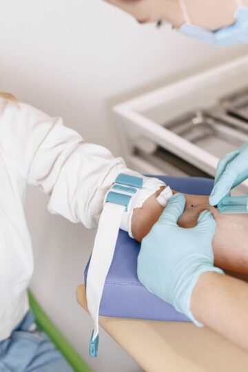 A healthcare worker in gloves prepares to draw blood from a patient's arm using a needle and tourniquet as part of a general healthcare routine in a medical setting.
