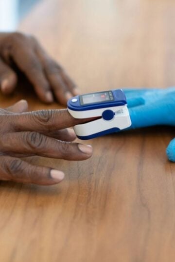 A person wearing a pulse oximeter on their finger while a gloved hand assists, both hands resting on a wooden surface.