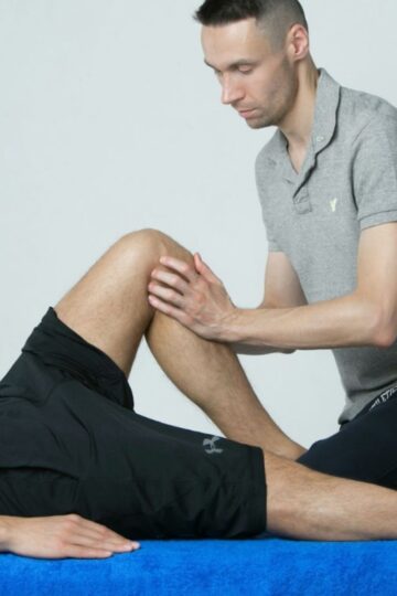 A man in a gray polo shirt performs a knee massage, often used by runners, on another person in black athletic shorts lying on a blue-covered table.