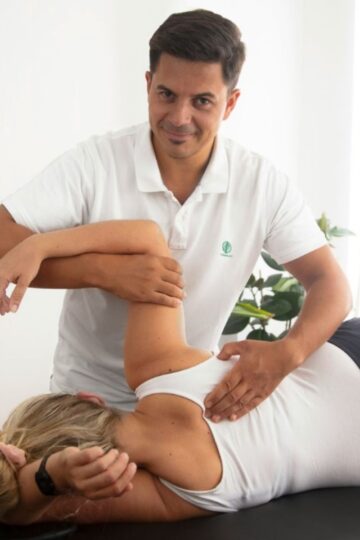 A physical therapist adjusts a woman's shoulder and back as she lies on her side on an examination table.