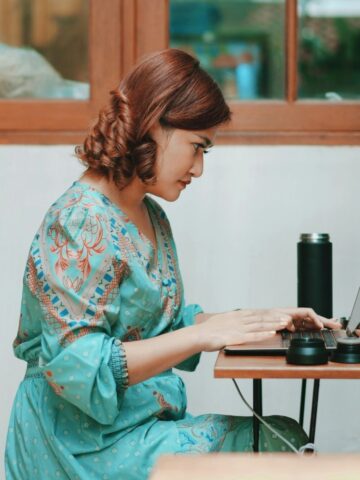 Woman in a blue patterned dress sits at a desk typing on a laptop, surrounded by a water bottle and small bottles-working indoors on her mom's guide to gaining credit with confidence.
