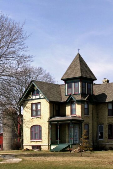 A large, two-story Victorian old house with a turret, yellow brick exterior, and green trim stands near a leafless tree and a red barn on a grassy lawn under a clear sky. Is this old house the one which is right for you?.
