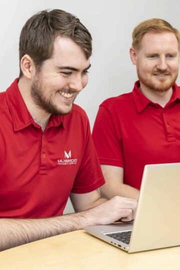 Two men in red polo shirts sit at a table, both looking at a laptop screen and smiling, as they discuss Expert IT Services for modern businesses.