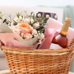 A wicker basket containing a bouquet of peach roses, a bottle of wine, and a box of chocolates, wrapped with pink paper and ribbon-perfect for thoughtful gifts to comfort in a box and support a loved one.