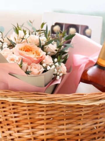 A wicker basket containing a bouquet of peach roses, a bottle of wine, and a box of chocolates, wrapped with pink paper and ribbon-perfect for thoughtful gifts to comfort in a box and support a loved one.