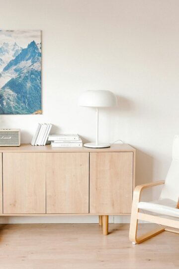 A light wood sideboard with books, a lamp, a radio, and decor stands against a white wall with a mountain photo; perfect for solving space dilemmas and making your home work. A chair with a brown pillow is nearby.
