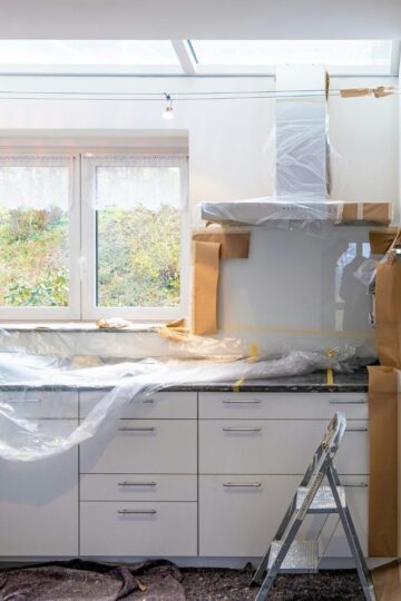A kitchen under renovation with cabinets, countertops, and appliances covered in protective plastic and paper; a step ladder is in front of the counter—a great example for those seeking home renovation tips.