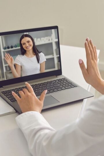 A person participates in an online treatment video call on a laptop, waving to another person displayed on the screen. A notebook and pen are on the desk, suggesting steps for managing bipolar disorder.