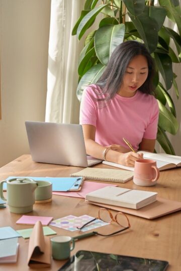 A woman sits at a wooden table with her laptop, notebooks, and mugs—organizing Uni Essentials and jotting down clever buys—near a large plant and window with sunlight streaming in.