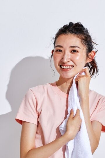 A young woman in a light pink shirt smiles while drying her face with a white towel against a plain white background, showing an easy post workout hair care routine to refresh hair after a workout without the need to wash.