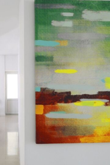 A colorful piece of Abstract wall art hangs on a white wall in a bright, minimal room with two chairs and another painting in the background—perfect inspiration to shop online for your own unique decor.