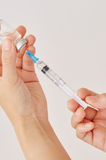 A person fills a syringe with liquid from a small glass vial, illustrating the preparation of GLP-1 drugs used in obesity care, set against a plain background.