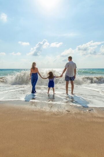 Three people, two adults and one child, stand hand in hand at the edge of the beach facing the ocean on a sunny day, embracing family health and wellness.