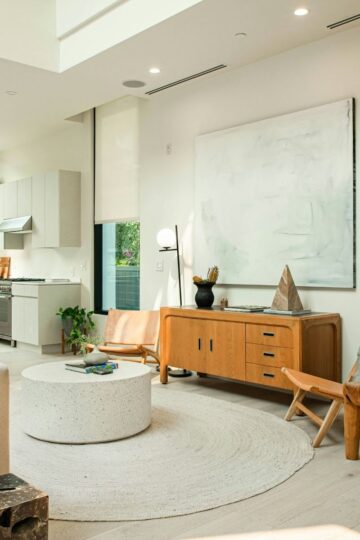 Modern living room with light wood furniture, large abstract artwork, round white rug, and open kitchen in the background. Natural light fills the space through large windows, enhancing comfort at home and overall home functionality.