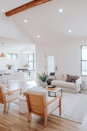A bright, modern living room with wooden floors, neutral furnishings, and large windows offers comfortable spaces, adjacent to an open kitchen with a breakfast bar and pendant lights.