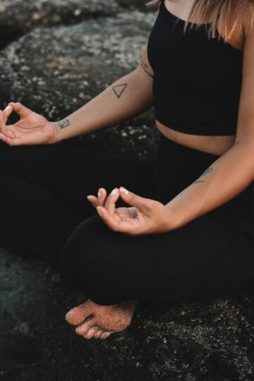 Person sitting cross-legged on rocks, wearing black workout clothes, with hands resting on knees in a meditation pose—embracing modern approaches to wellness.