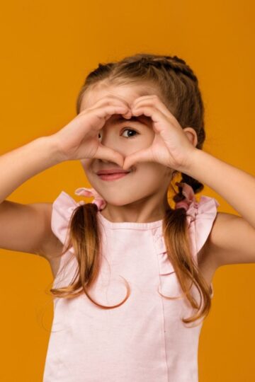 Young girl with braided hair in a pink dress makes a heart shape with her hands over her eyes against an orange background, capturing the pure joy of giving and kindness.