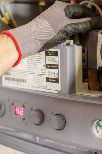 A person wearing gloves adjusts controls on a gas boiler, with a digital display reading 42 and various buttons, labels, and parts visible.