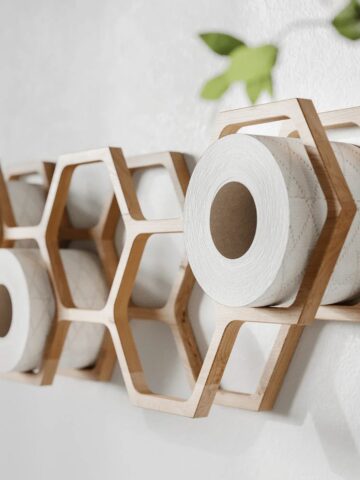 Wall-mounted wooden hexagonal shelves holding rolls of toilet paper showcase smart bathroom organisation, with part of a green plant visible in the corner.