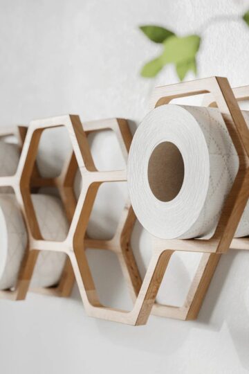 Wall-mounted wooden hexagonal shelves holding rolls of toilet paper showcase smart bathroom organisation, with part of a green plant visible in the corner.