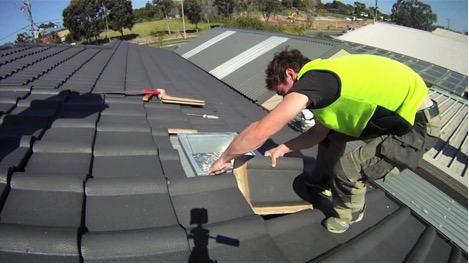 A person wearing a yellow safety vest installs a skylight or vent on a tiled roof, using tools and materials placed nearby.