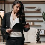 A woman in business attire stands in an office, holding a clipboard and pen. A statue of Lady Justice is visible on the desk beside her, reflecting her role as a trusted family solicitor in Scotland.
