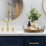 A bathroom countertop with gold faucet fixtures, a round mirror, and a tray holding a vase with greenery, soap, lotion, and a candle.