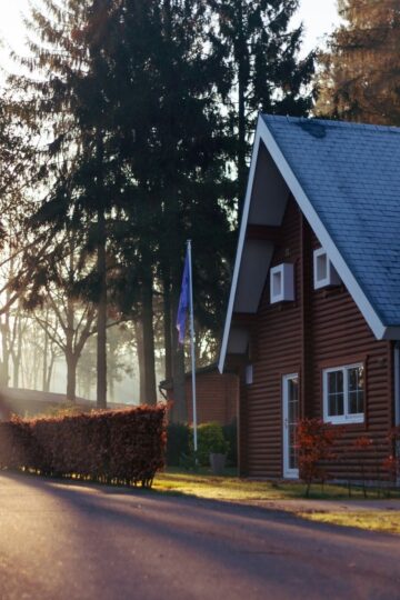 A wooden house with a steep roof sits beside a paved road, surrounded by trees and morning sunlight, offering age-friendly home features and excellent foundation stability.