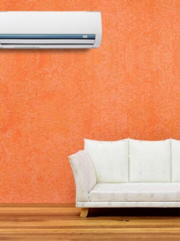 A white wall-mounted air conditioner, perfect for easy AC maintenance, is installed above a white sofa set against an orange textured wall and wooden floor.
